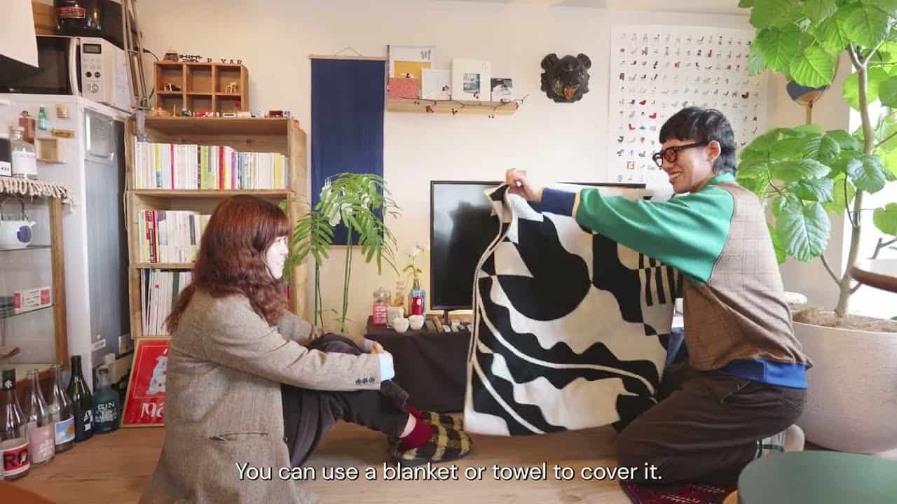 Ryu kneeling in the Yoshinani living room, holding a black-and-white patterned blanket up to cover the television, Kamado seated on the floor