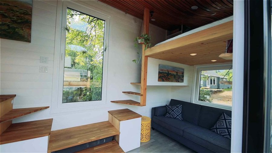 Living room with gray sofa, floating wooden stairs and large windows in the curved-roof tiny house