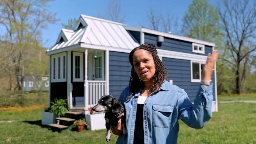 Woman holding a dog in front of a modern tiny house on a sunny day.